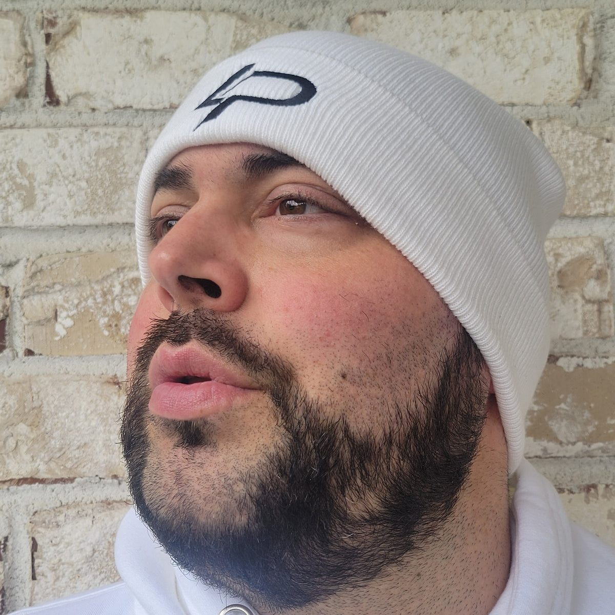 Bearded man looking upward wearing a white ribbed knit beanie with a dark embroidered LP logo, against a stone brick wall. Beanie apparel.