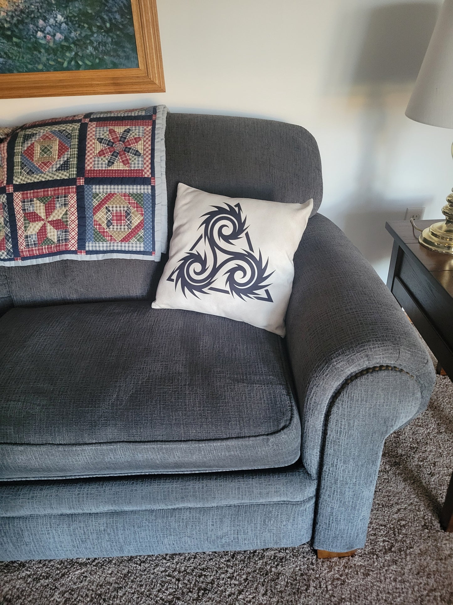Beige throw pillow featuring a black Norse triskelion with spiral arms, displayed on the right corner of a gray sofa — throw pillow.