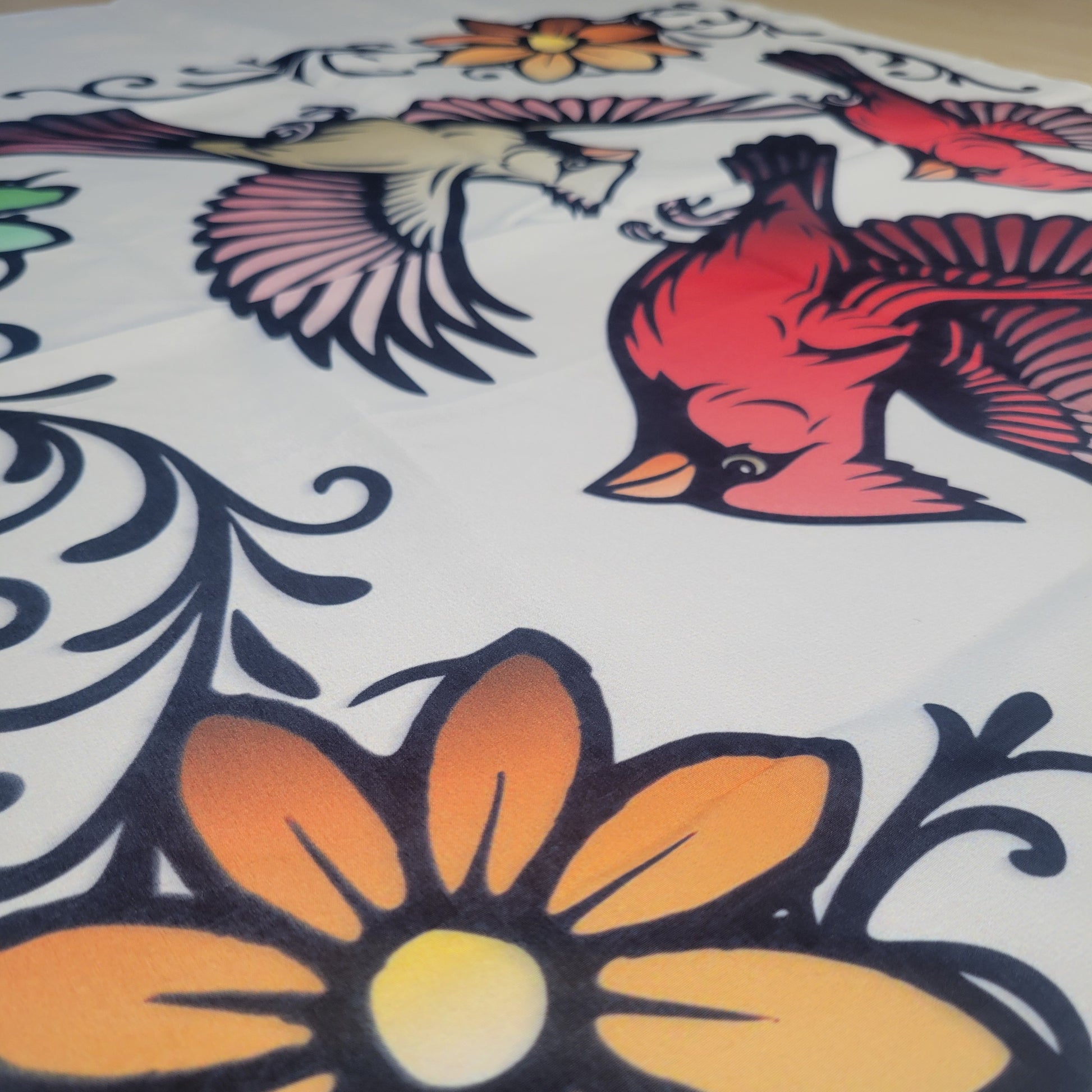 Bold tattoo-style illustration of a red cardinal and pale bird in flight surrounded by orange flowers and black swirling vines on a beige fabric tapestry.