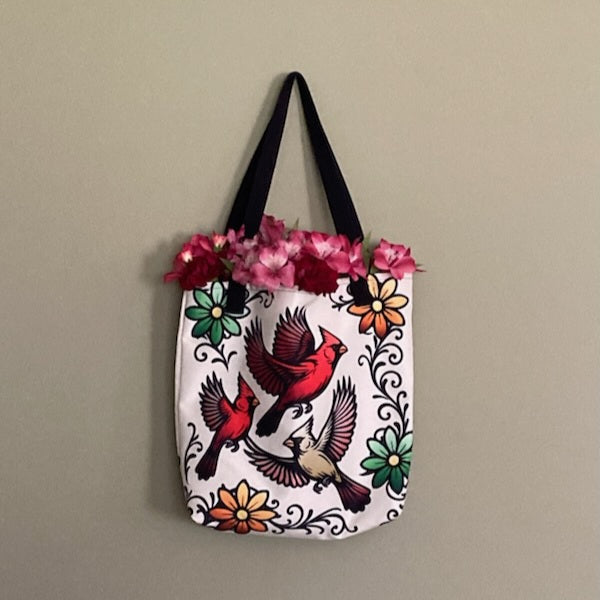 Beige tote bag with black handles featuring three cardinals in flight surrounded by colorful folk-art flowers and dark swirling vines, displayed hanging on wall with pink flowers tucked inside.