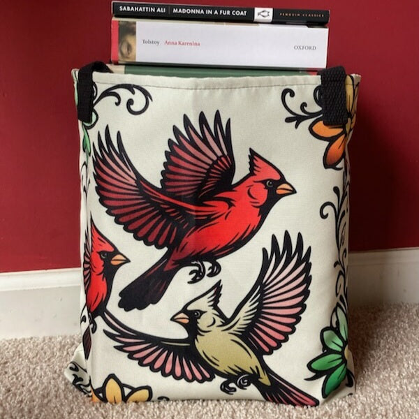Cream fabric tote bag printed with bold tattoo-style flying cardinals in red and gold, with floral accents, holding books upright.