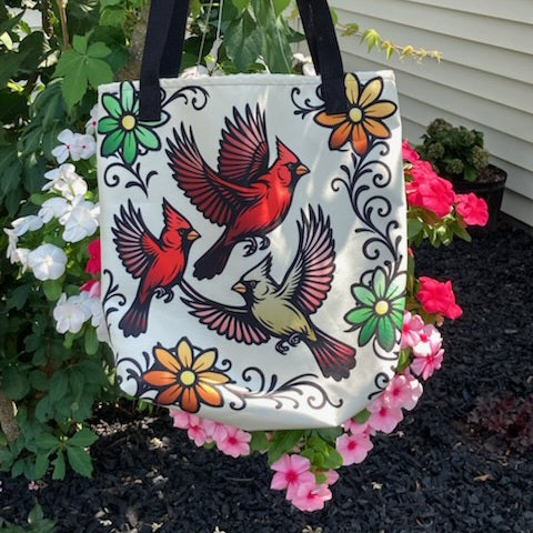 Cream tote bag with black handles featuring bold illustrated red cardinals in flight surrounded by decorative floral scrollwork in red, orange, and green, hanging outdoors among pink flowers.