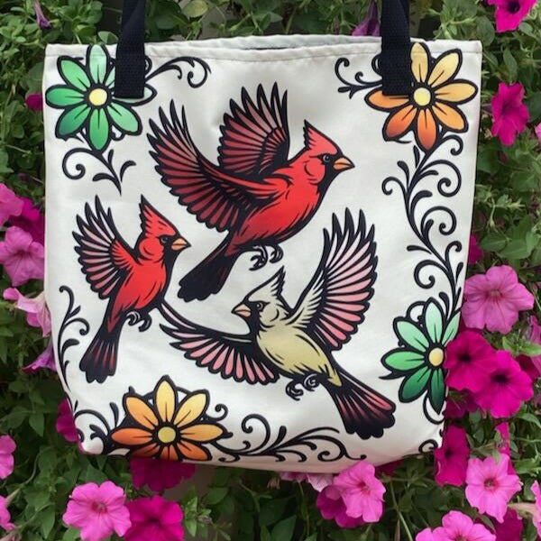 Three red and cream cardinals in flight surrounded by bold floral vines on a cream canvas tote bag with black straps. Photographed on a pink flower bed.