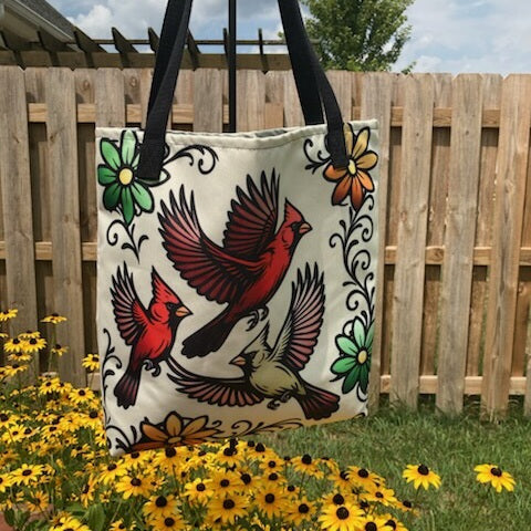 Three flying cardinals in bold tattoo-style art with colorful flowers on a cream canvas tote bag with black handles. Hanging over a bed of yellow and black flowers.
