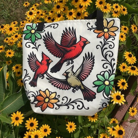 Cream canvas tote bag printed with three flying cardinals and decorative floral scrollwork in bold tattoo-style illustration, displayed among yellow wildflowers.