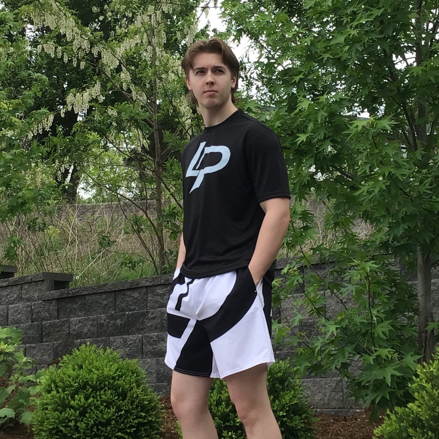 Male model outdoors wearing a black t-shirt with white "LP" logo graphic and bold black-and-white printed athletic shorts, surrounded by green trees and a stone wall. Apparel lifestyle photo.