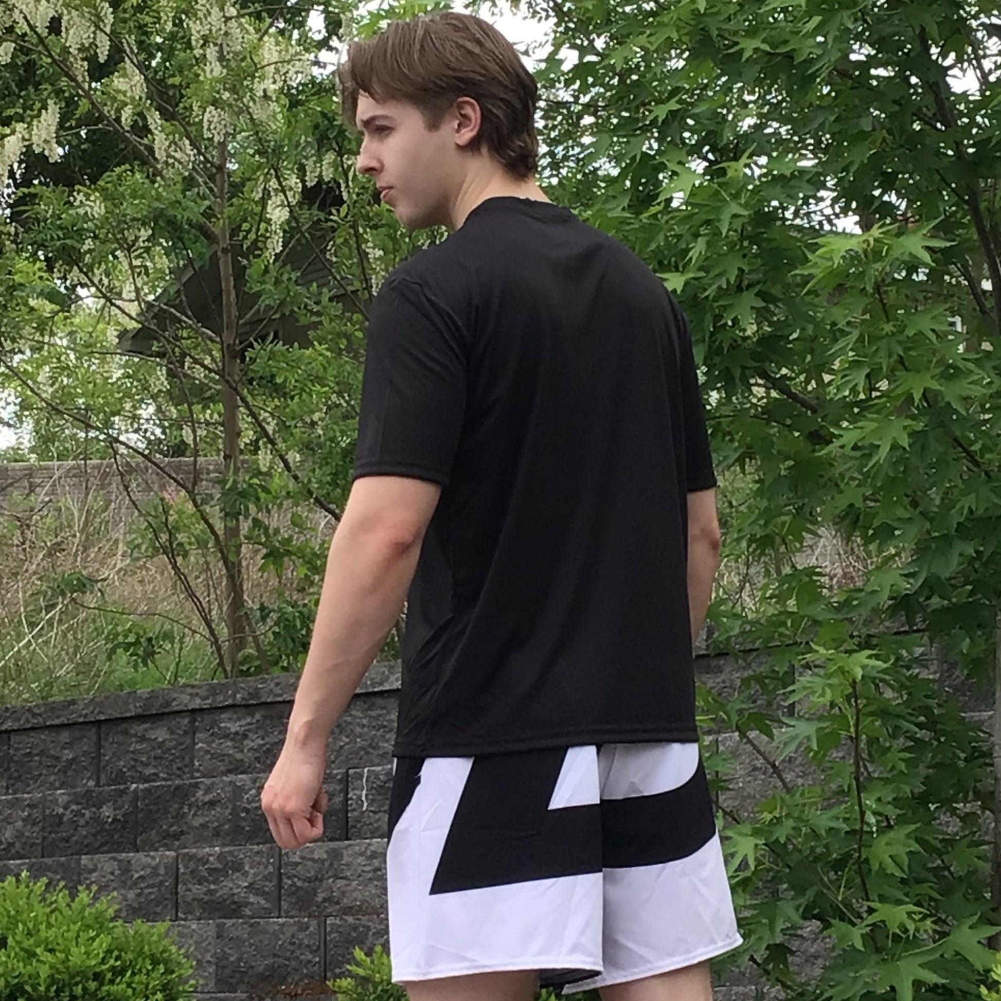 Male model shown from the back left side wearing a plain black short-sleeve t-shirt and white athletic shorts with bold black graphic, outdoors against green foliage. Apparel lifestyle photo.