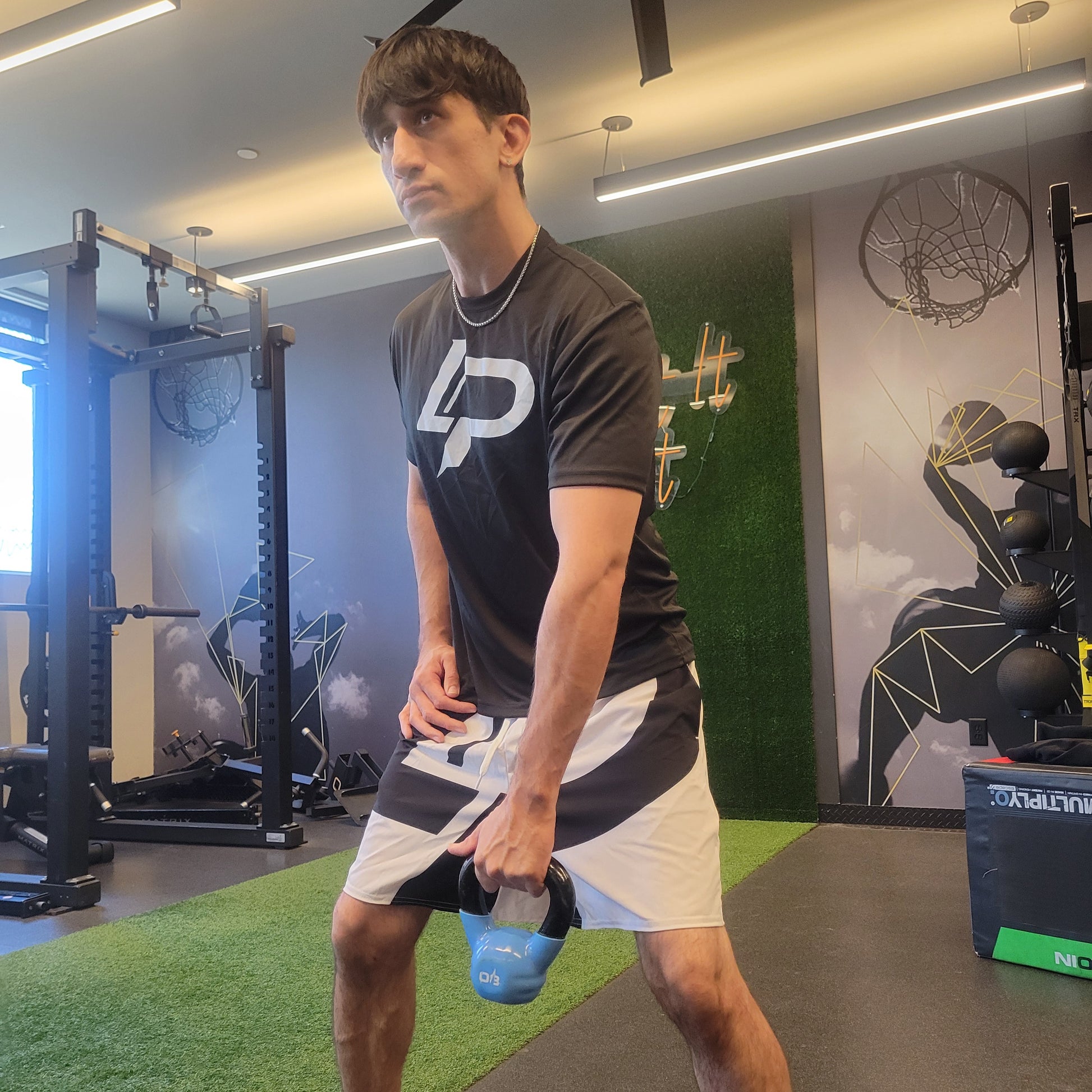 Man holding a blue kettlebell in a modern gym, wearing a black LP graphic tee and white LP printed athletic shorts, apparel lifestyle photo.