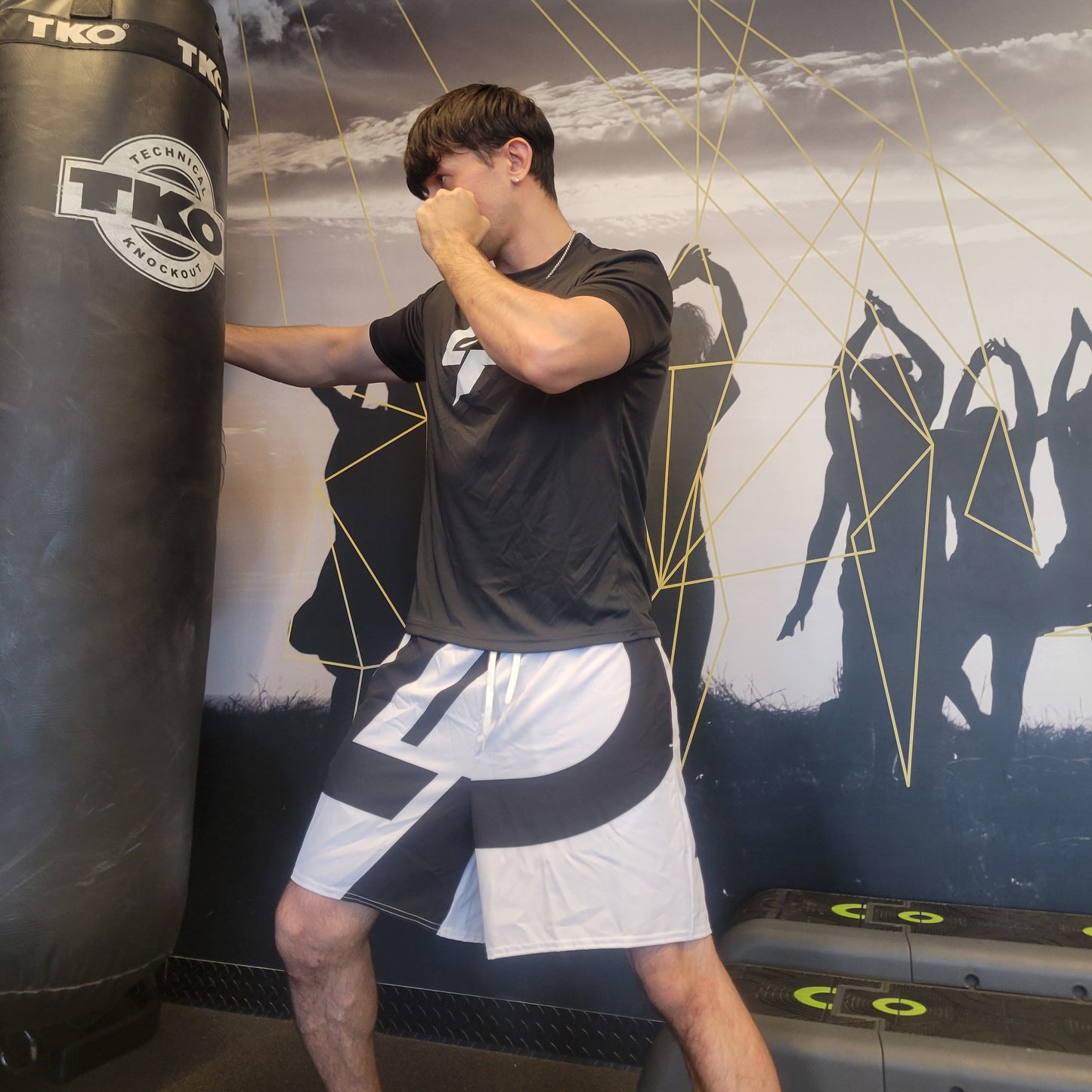 Male model striking a TKO punching bag wearing a black LP graphic tee and bold black-and-white athletic shorts with large LP logo print, shot in a gym setting. Apparel lifestyle photo.