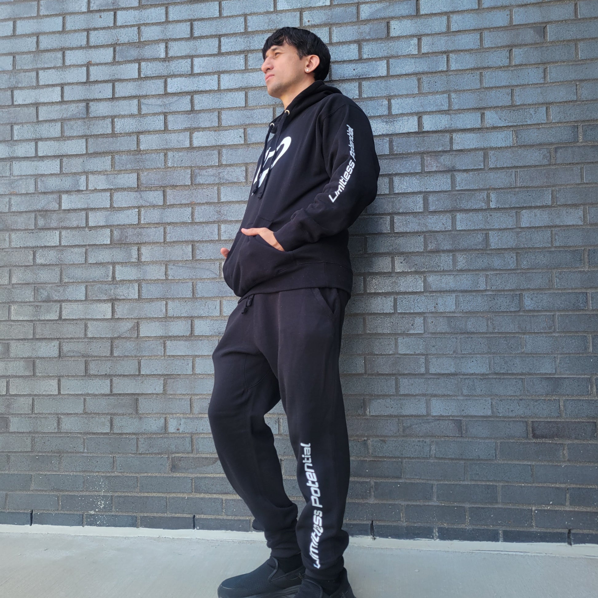 Man leaning against a gray brick wall wearing a black hoodie and joggers set with white "Limitless Potential" text on the left sleeve and left pant leg, apparel lifestyle photo.