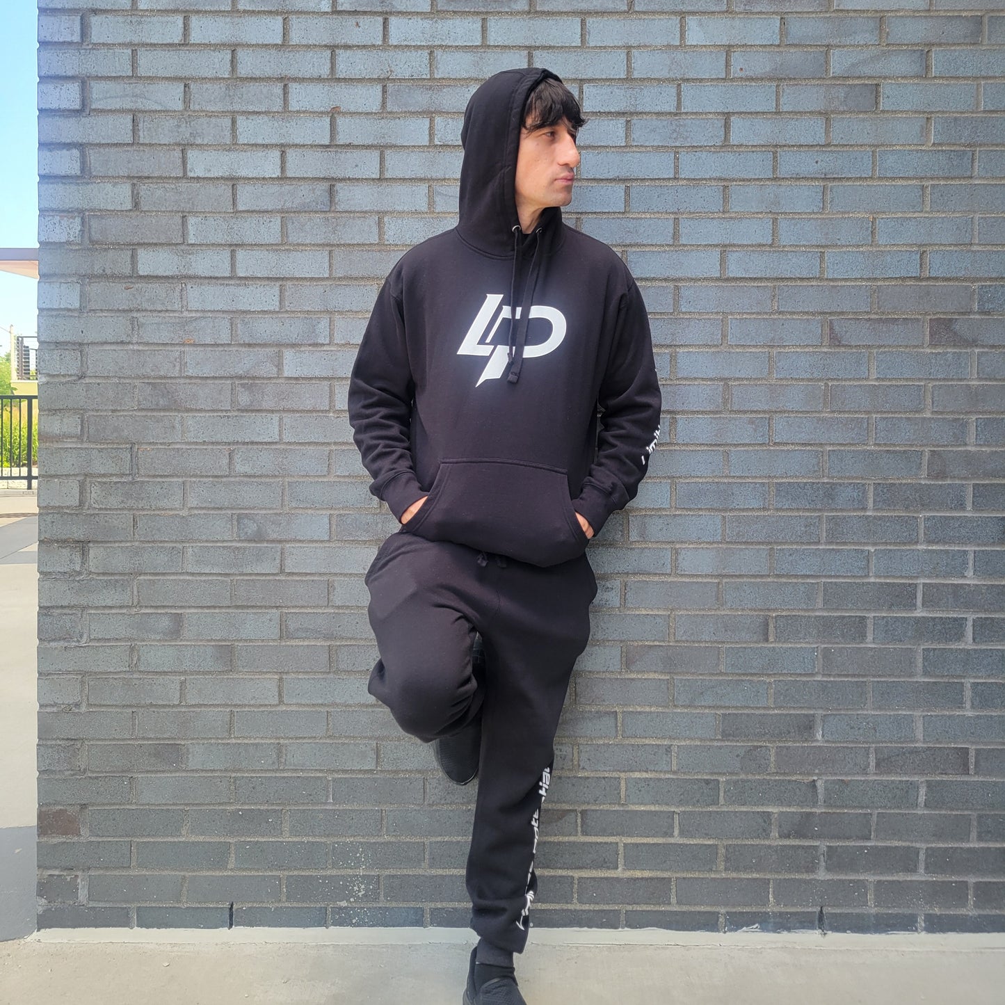 Man leaning against a gray brick wall wearing an all-black hoodie and jogger set with a bold white "LP" logo on the chest and white text along the left sleeve and pant leg, apparel lifestyle photo.