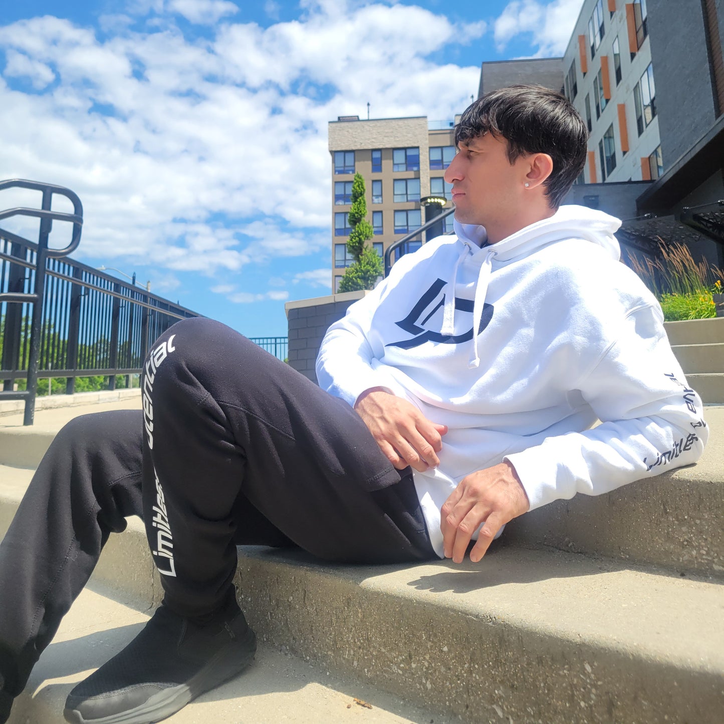 Male model seated on outdoor concrete steps wearing a white hoodie with a black logo graphic and black jogger pants with "Limitless" text along the leg, urban background, apparel lifestyle photo.