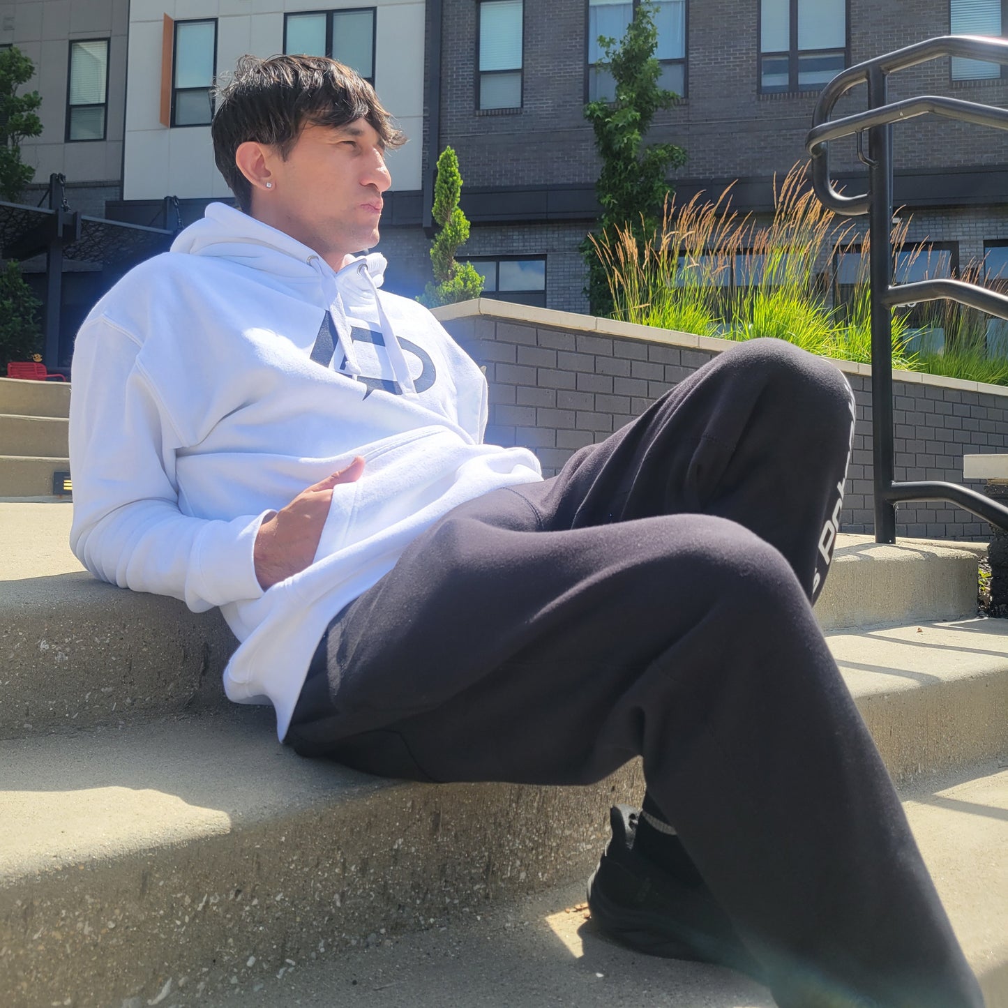 Male model seated on outdoor concrete steps wearing a white LP graphic hoodie and black sweatpants, looking to the side in natural sunlight. Apparel lifestyle photo.