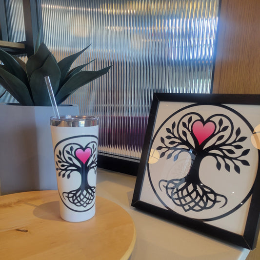 White tumbler and matching, black-framed print featuring a black Yggdrasil tree-of-life design with a pink heart, displayed together on a table with potted plant.