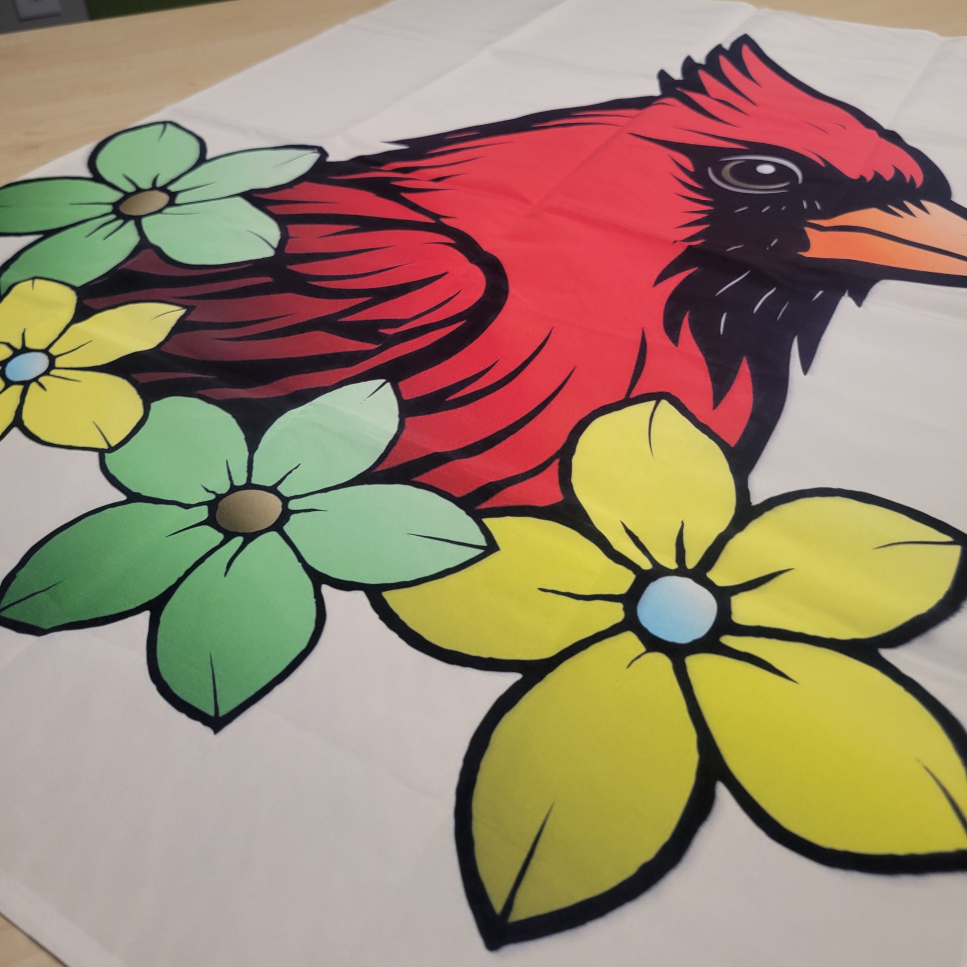 Bold tattoo-style graphic of a red cardinal surrounded by green and yellow flowers with black outlines on a beige fabric tapestry.