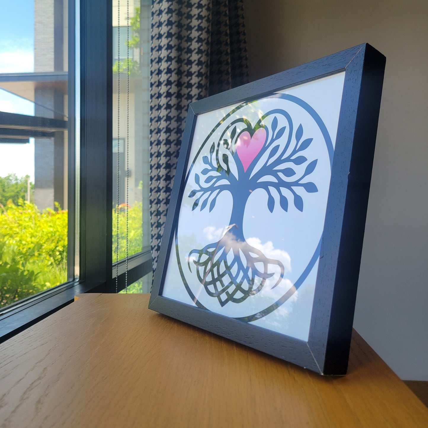 Black framed wall art featuring a Norse Tree of Life with interwoven roots, leafy branches, and a pink heart at center, displayed on a wooden surface near a sunlit window.