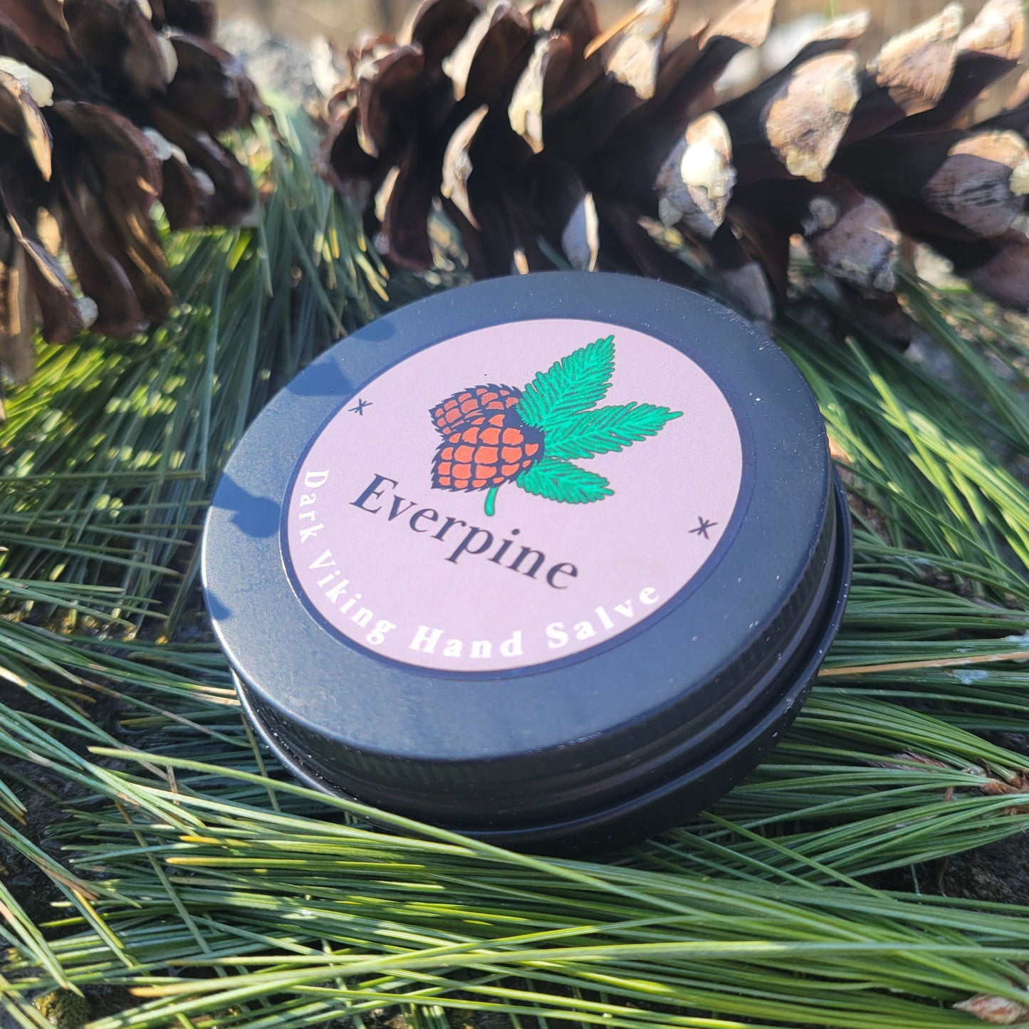 Black metal tin of Everpine Dark Viking Hand Salve, its mauve label featuring a illustrated pine cone and green pine branches, nestled among real pine needles and pine cones outdoors.
