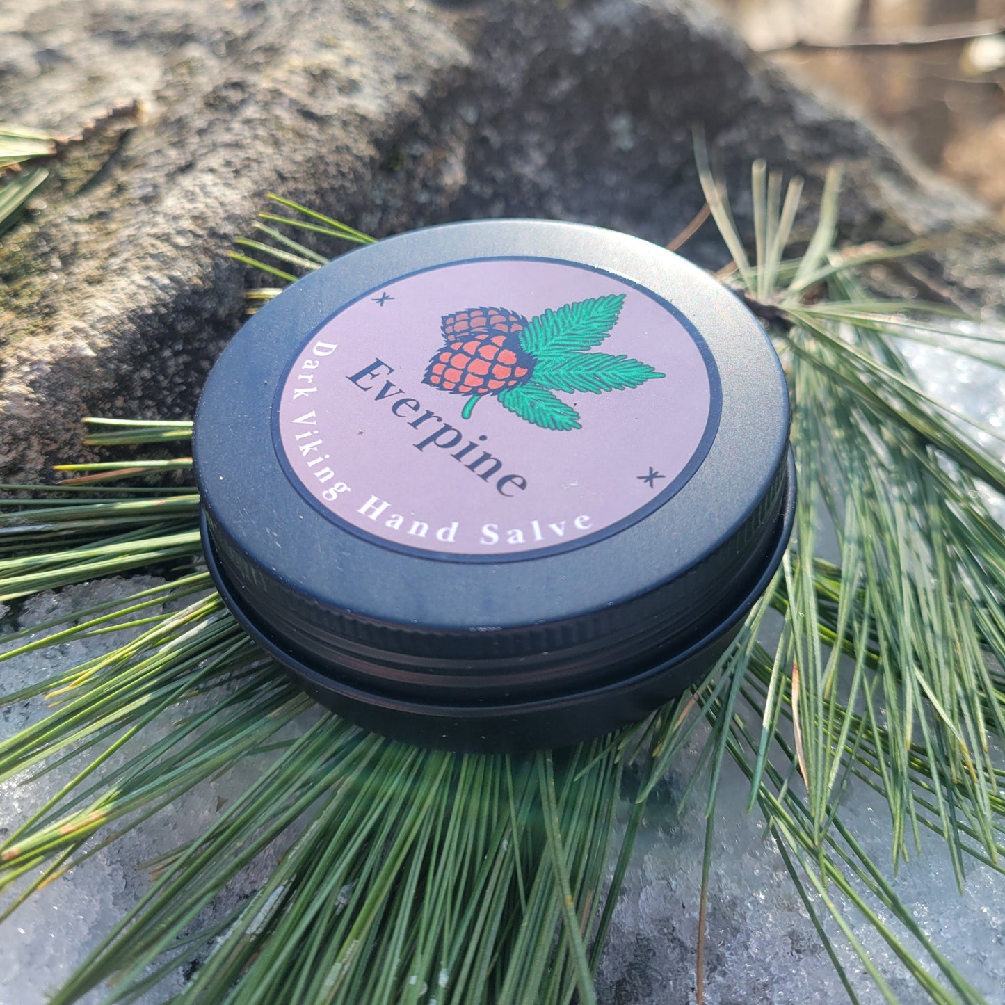 Black metal tin labeled "Everpine Dark Viking Hand Salve" with a mauve circular label featuring a pine cone and green branches, resting on snow surrounded by fresh pine needles — handmade hand salve.