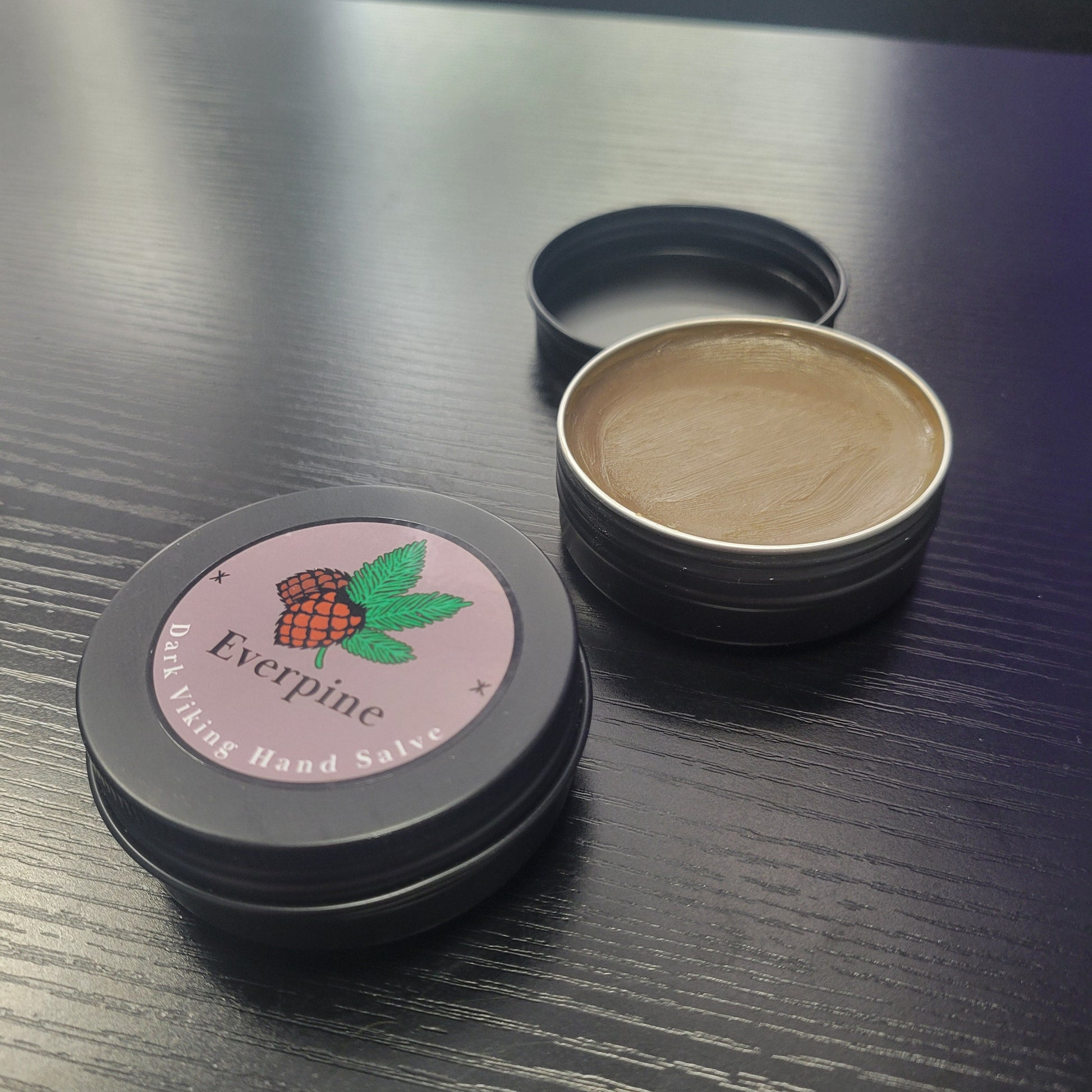 Two small black metal tins on a dark wood surface — one closed with a mauve label reading "Everpine Dark Viking Hand Salve" with a pine illustrations, one open revealing the dark  brown salve inside.