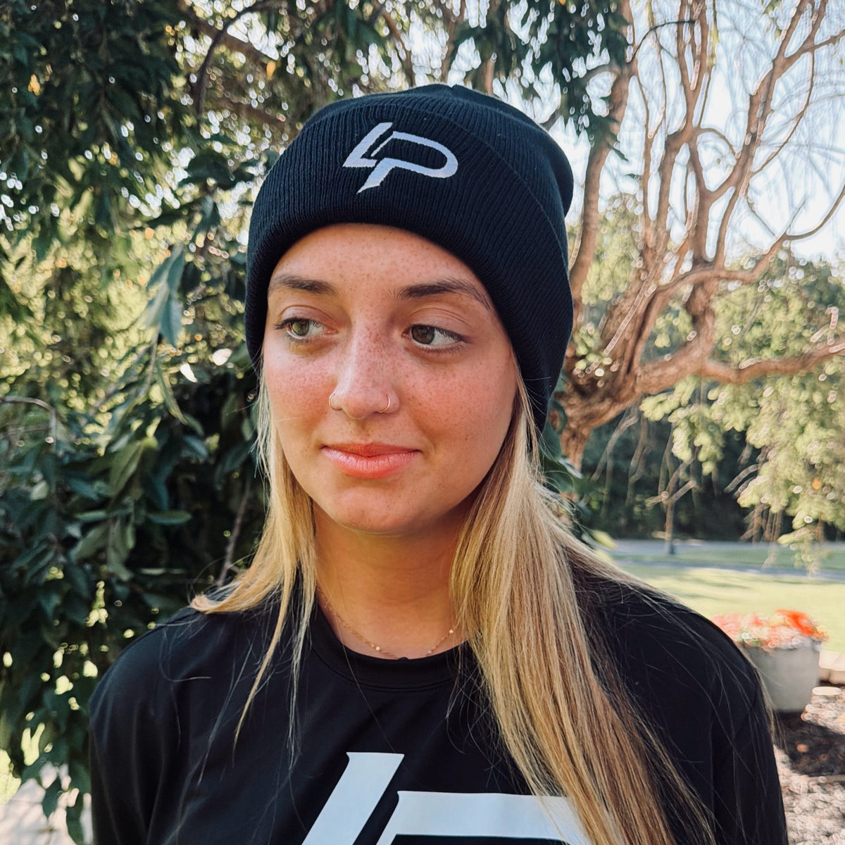 Model wearing a black knit beanie with a gray embroidered "LP" logo, paired with a matching black graphic tee, outdoors with trees in the background. Beanie lifestyle product photo.