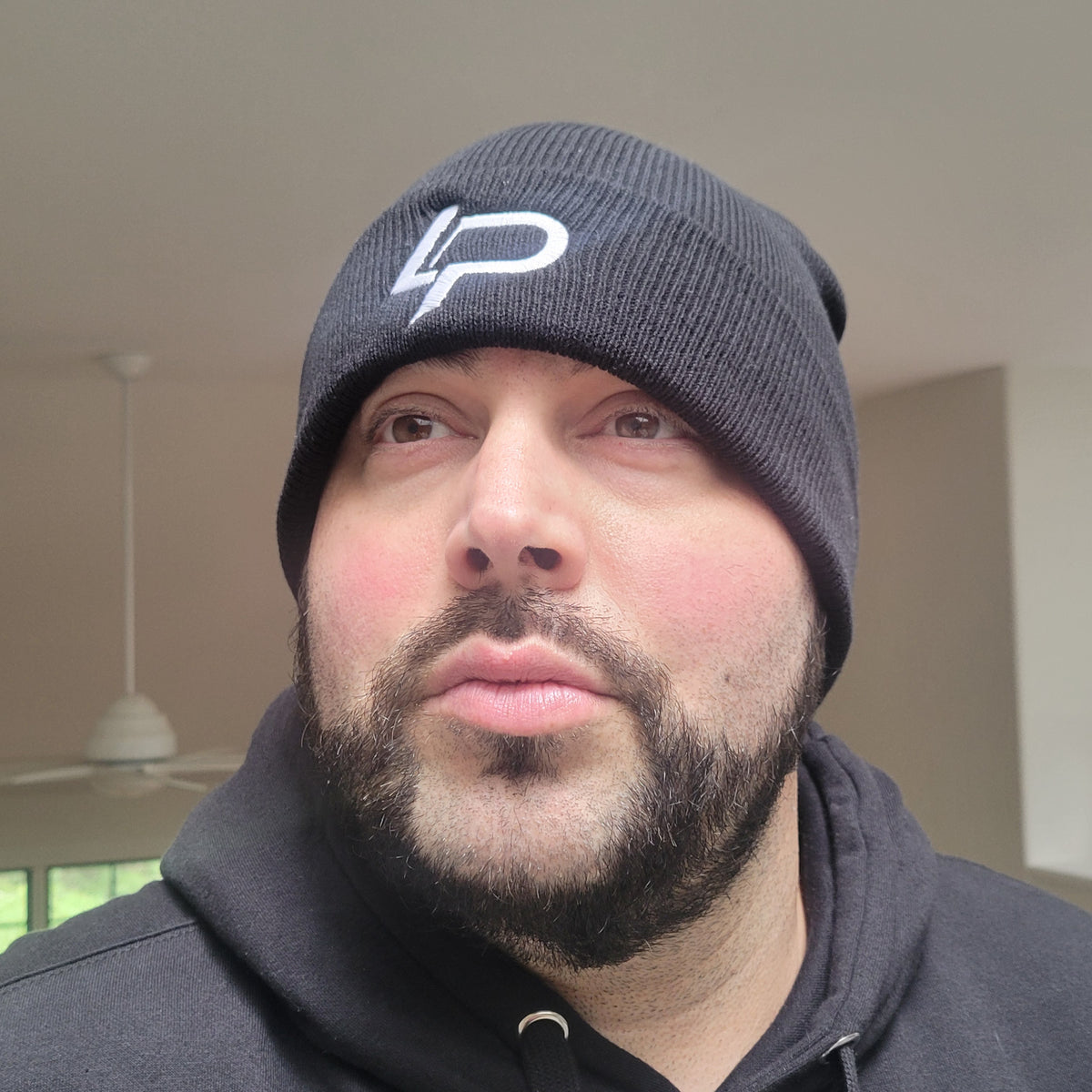 Black ribbed beanie with white embroidered LP logo worn by a bearded male model, paired with a black hoodie, lifestyle product photo — beanie apparel.