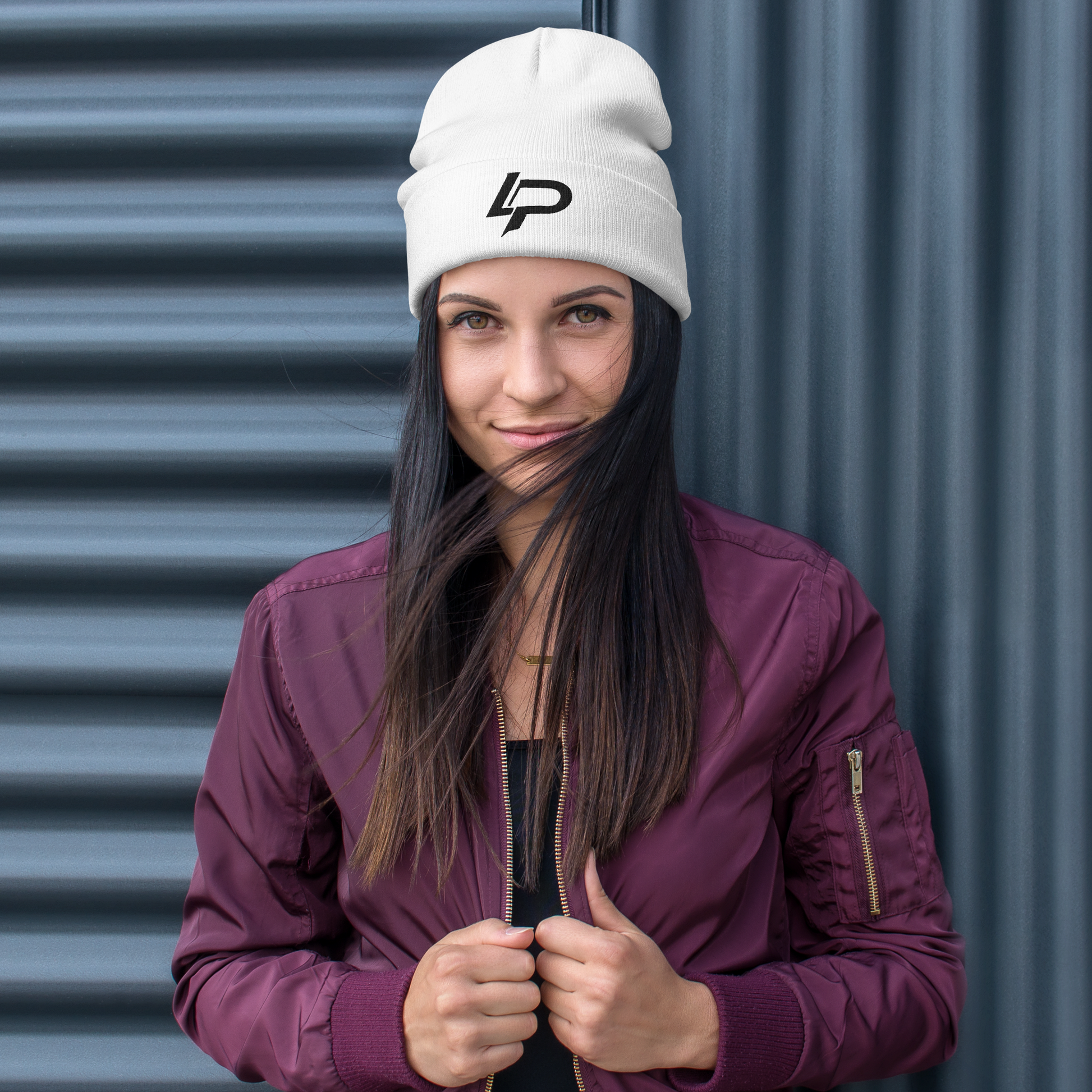 Woman wearing a white cuffed beanie with a bold black "LP" logo embroidered on the front, paired with a burgundy bomber jacket against a corrugated metal backdrop. Embroidered beanie.