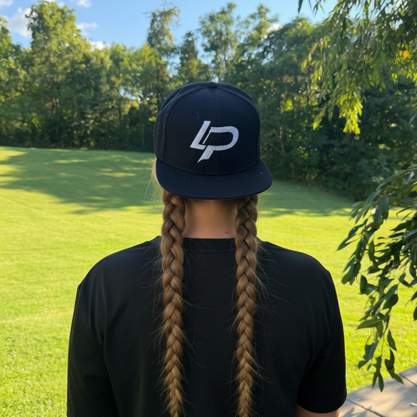 Person seen from behind wearing a black snapback hat with a bold white "LP" logo, with long braided hair, standing outdoors in a green landscape; snapback cap apparel.