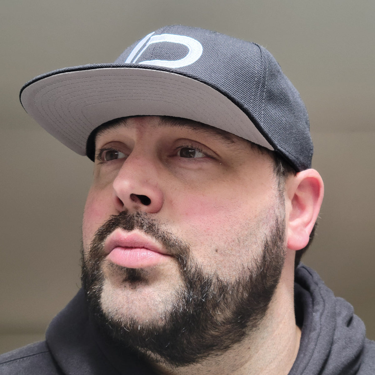 Bearded male model wearing a black snapback hat with a preview of the "LP" logo embroidered in white. Apparel and headwear lifestyle photo.