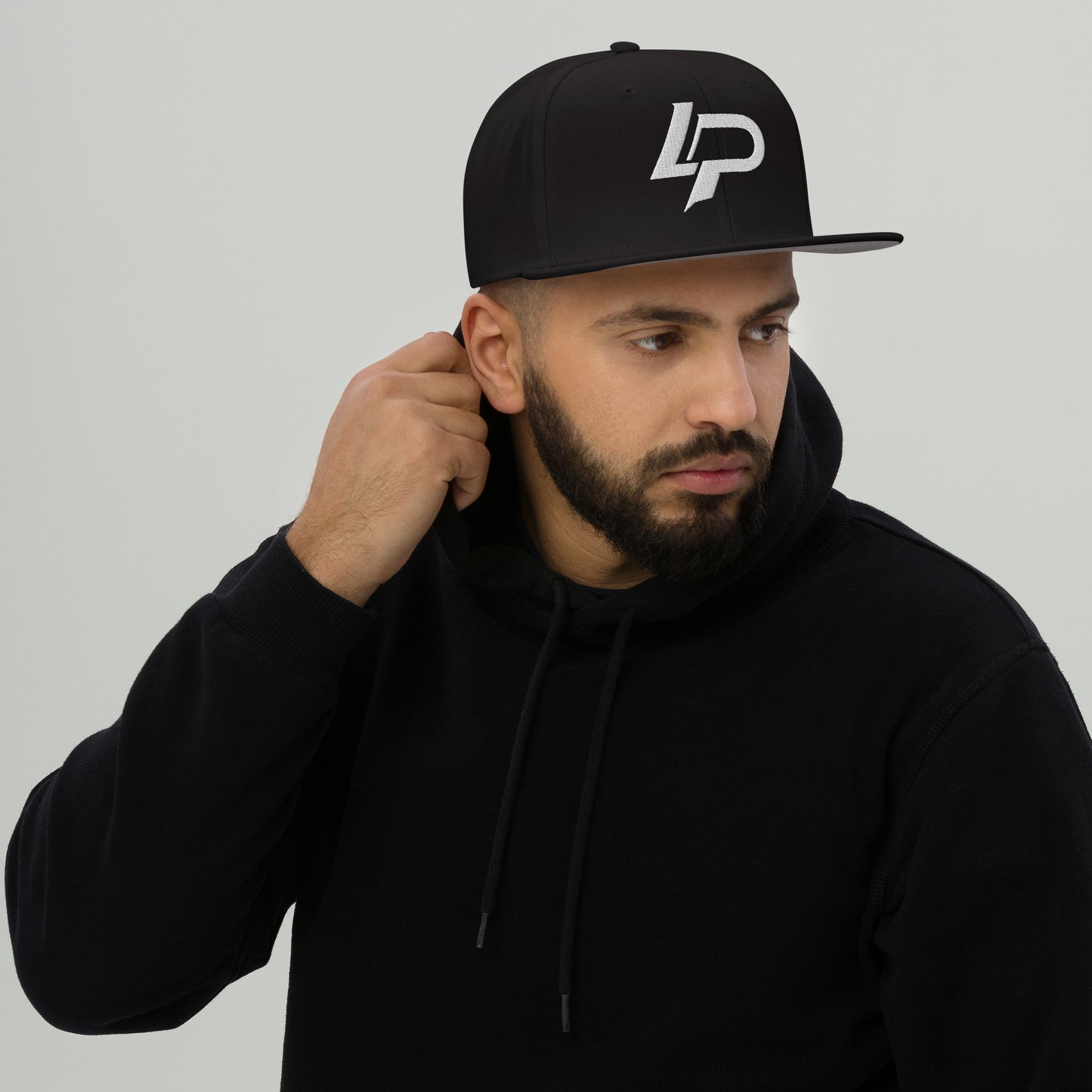 Man wearing a black flat-brim snapback hat with a white embroidered "LP" logo, with a black hoodie, against a light gray background. Snapback hat.