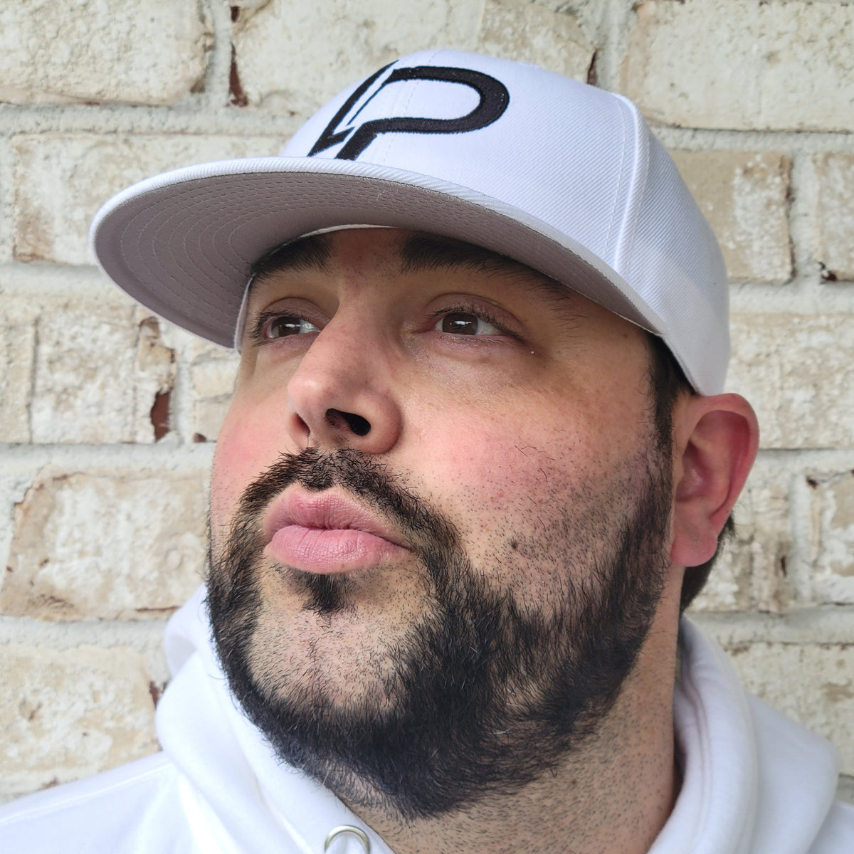 Bearded man looking upward wearing a white snapback cap with black embroidered LP logo, paired with a white hoodie, against a brick wall background. Snapback hat lifestyle photo.