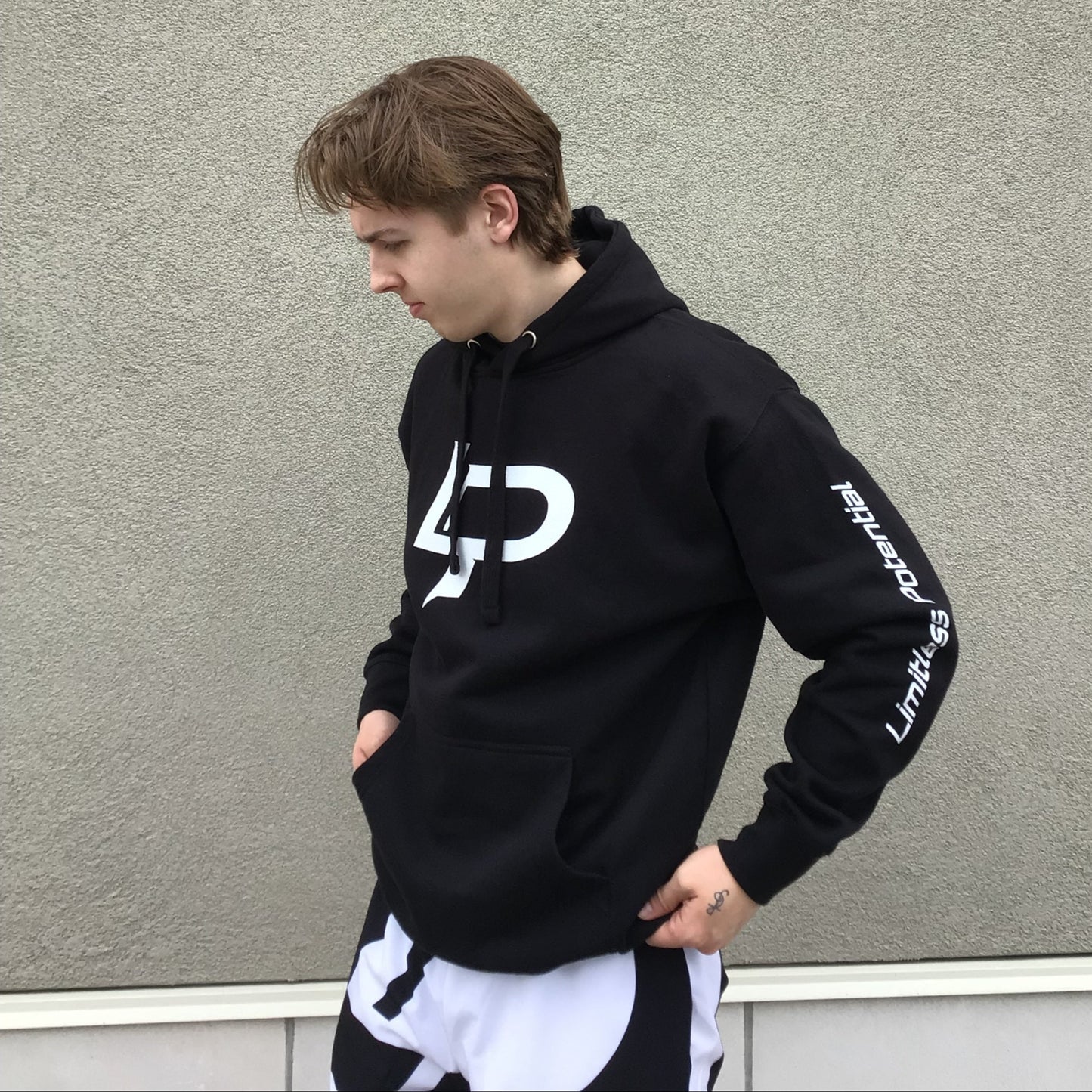 Photograph of a model wearing a black hoodie with white "LP" logo on chest and "Limitless Potential" text sown the left sleeve against a grey wall.