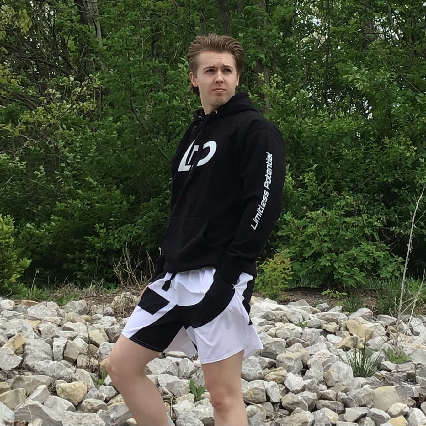 Young man modeling a black hoodie with a white "LP" logo and "Limitless Potential" sleeve text, paired with black-and-white shorts, standing on rocks outdoors — apparel lifestyle photo.