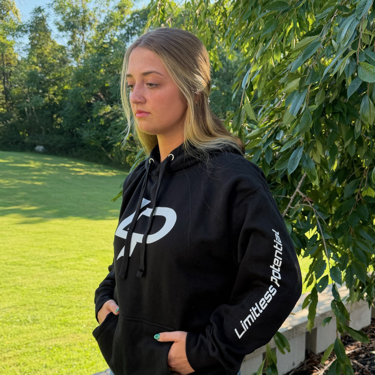 Black pullover hoodie with large white LP logo on chest and "Limitless Potential" printed along the sleeve, modeled outdoors against a green garden backdrop. Apparel.