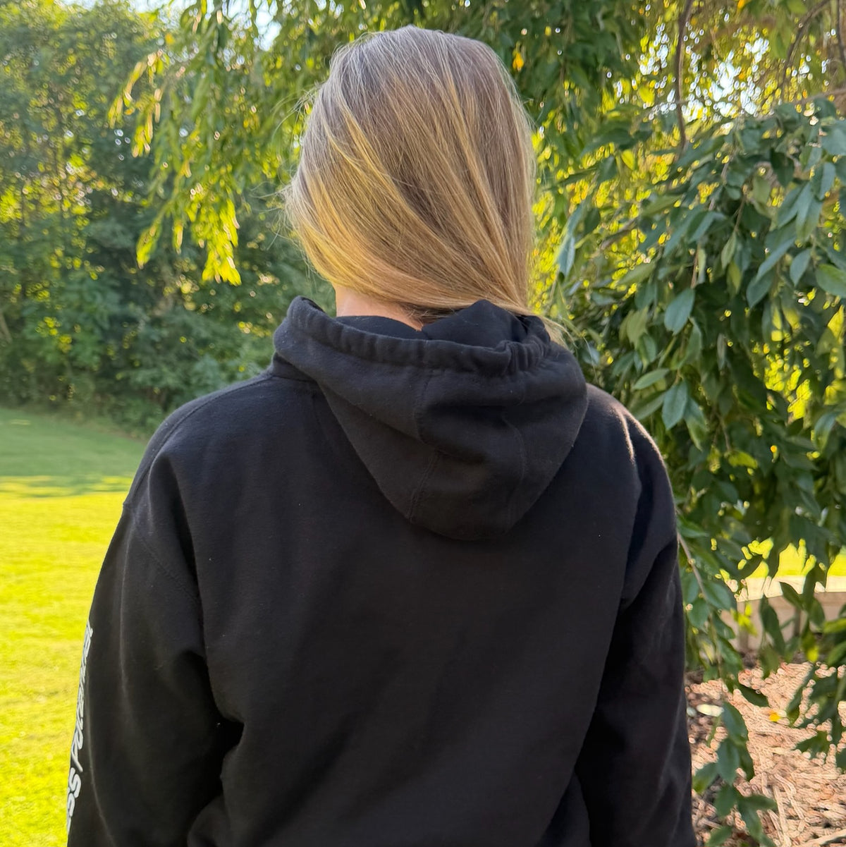 Back view of a female model with blonde hair wearing a black pullover hoodie, with white text on the left sleeve, photographed outdoors among green trees. Apparel.