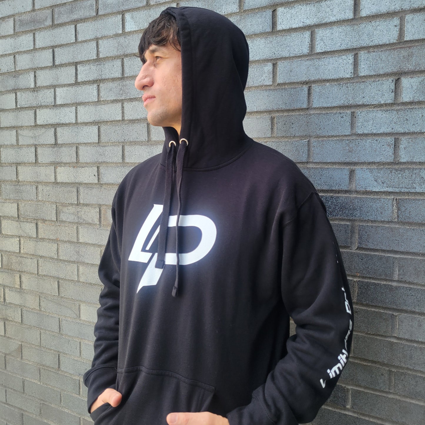 Man wearing a black pullover hoodie with a bold white "LP" logo on the chest and white sleeve text, photographed against a gray brick wall. Apparel lifestyle photo.
