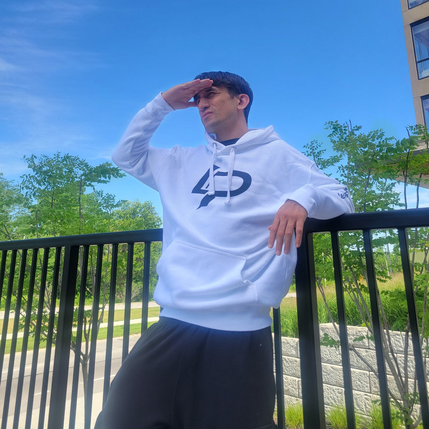 Man wearing a white pullover hoodie with a bold black LP logo graphic, shielding his eyes from the sun outdoors against a bright blue sky and green trees; hoodie lifestyle apparel.