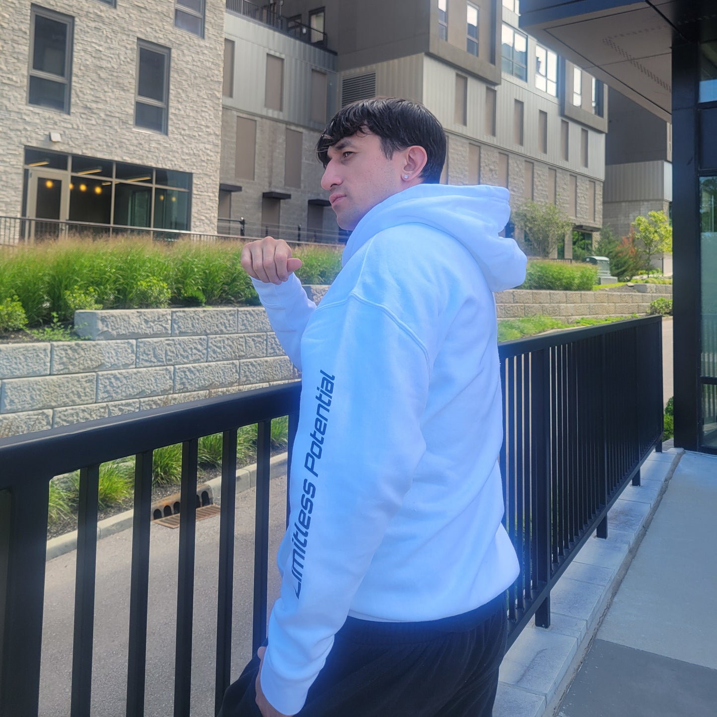 Man posing outdoors in a white pullover hoodie with "Limitless Potential" printed vertically along the sleeve in black, set leaning forward against an urban backdrop. Apparel lifestyle photo.