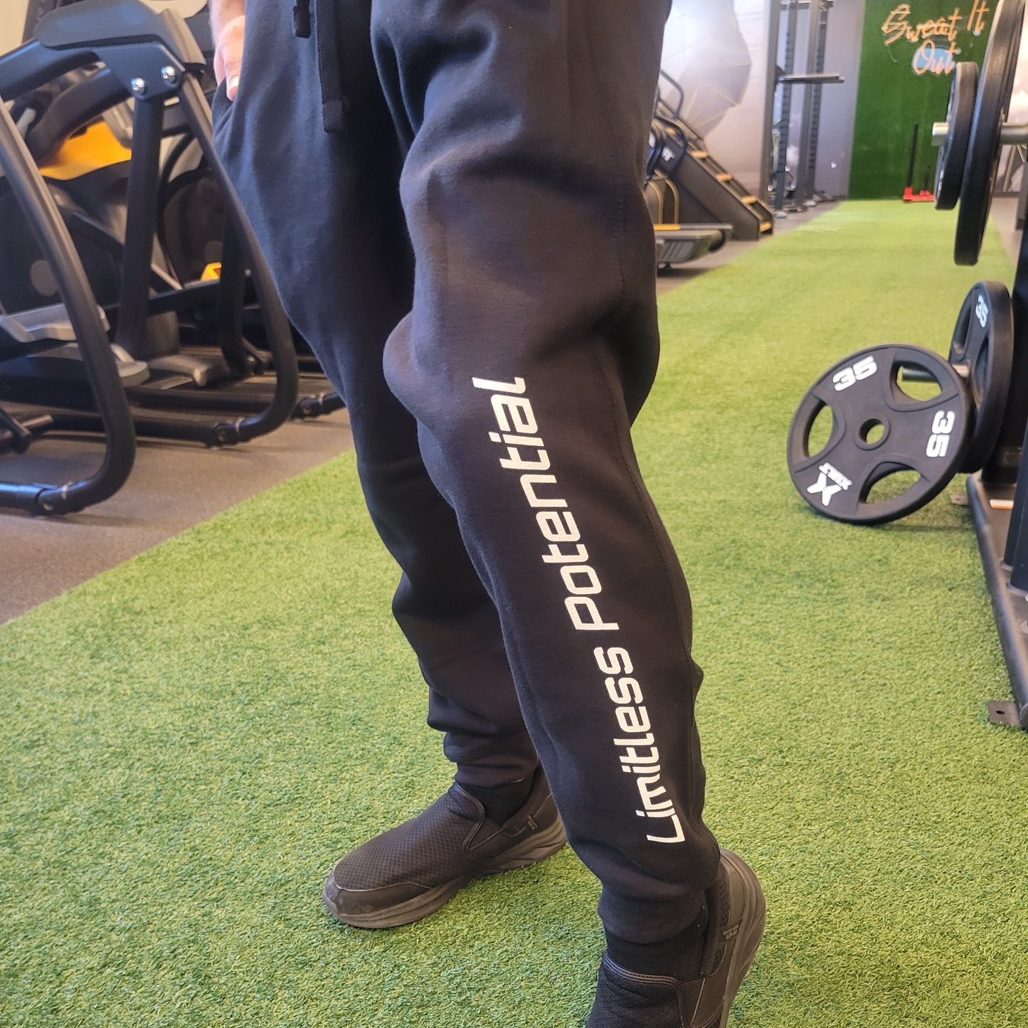 Black jogger sweatpants with "Limitless Potential" printed vertically in white text along the left leg, worn in a gym setting, apparel.