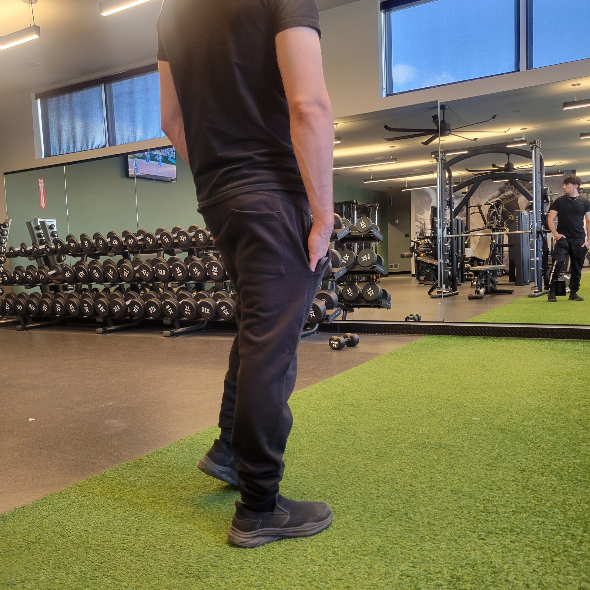 Person wearing all-black joggers and t-shirt standing in a modern gym with dumbbell racks and green turf flooring; Shown from the back. apparel lifestyle photo.
