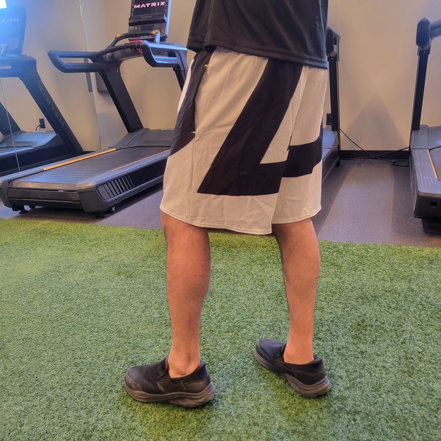 White athletic shorts with a bold black diagonal graphic, worn by a model in a gym setting, shown from behind. Apparel.