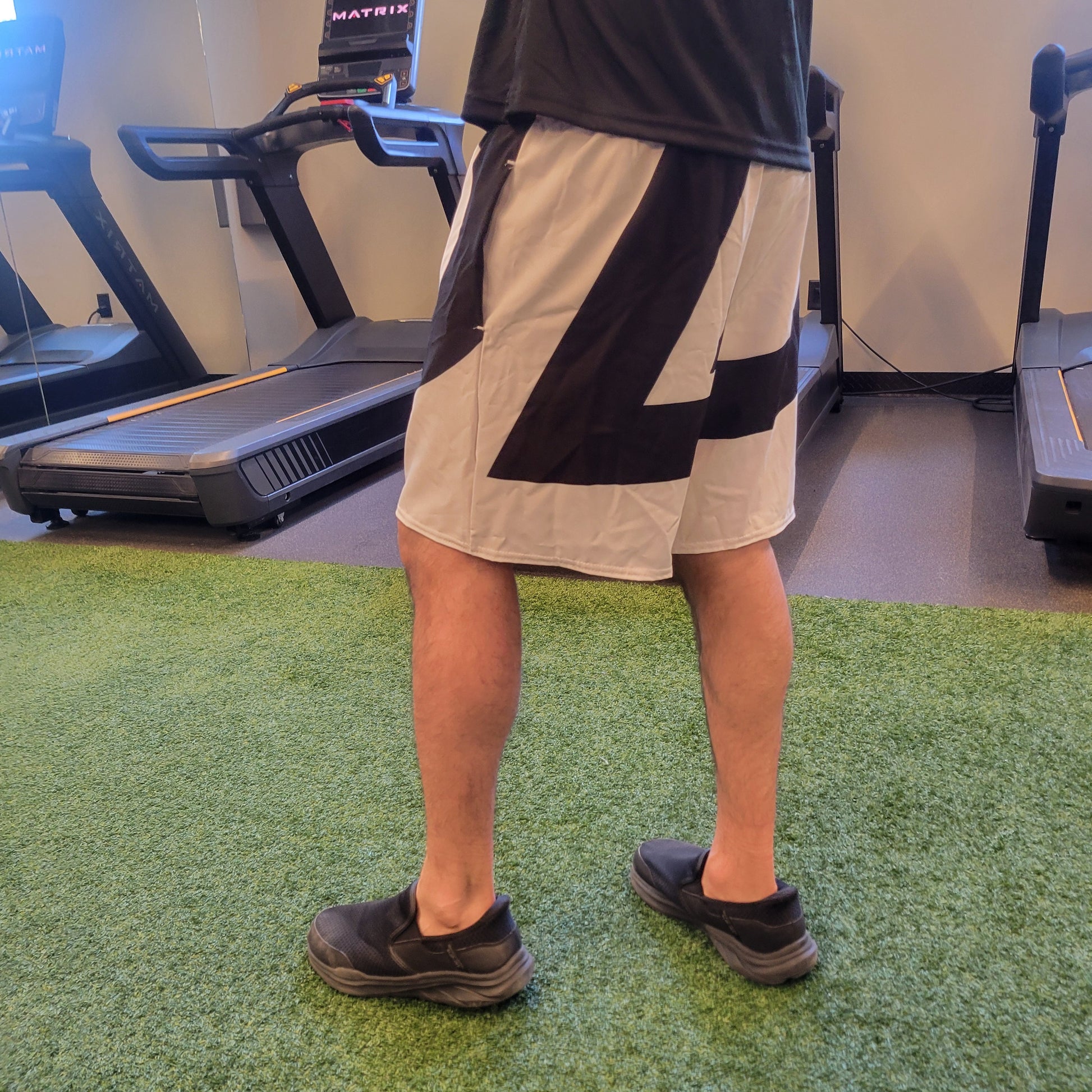 White athletic shorts with a bold black diagonal graphic, worn by a model in a gym setting, shown from behind. Apparel.