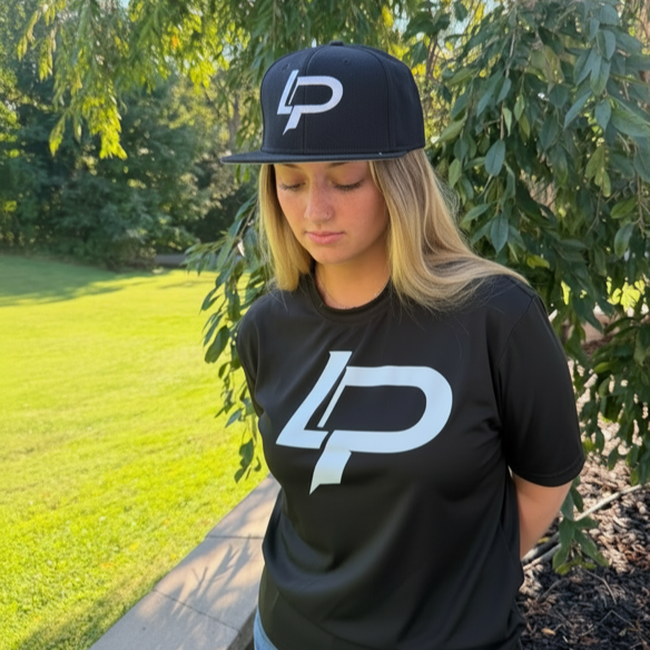 Young woman wearing a black snapback hat and matching black tee, both featuring a bold white "LP" logo, posed outdoors in natural light. Apparel lifestyle photo.