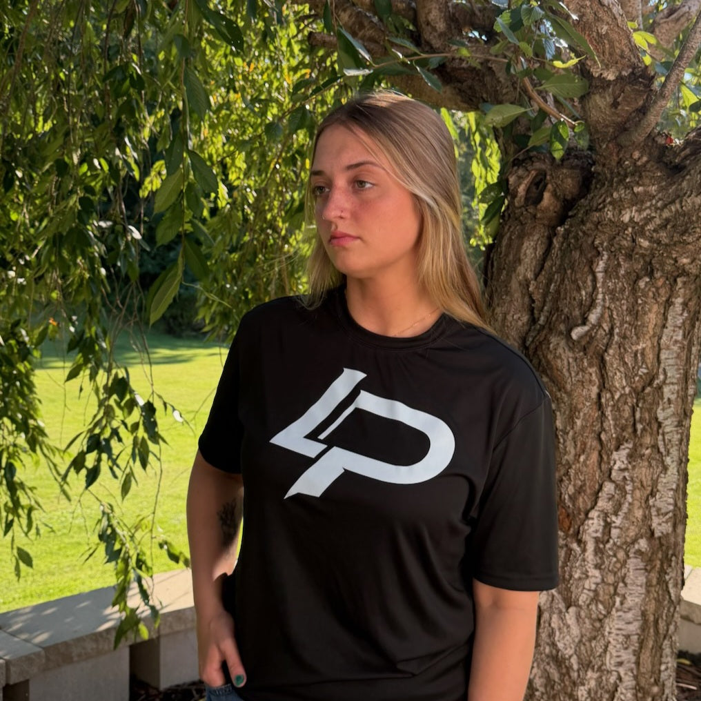 Woman wearing a black crew-neck t-shirt with a bold white "LP" logo graphic, standing outdoors beside a tree. Apparel.