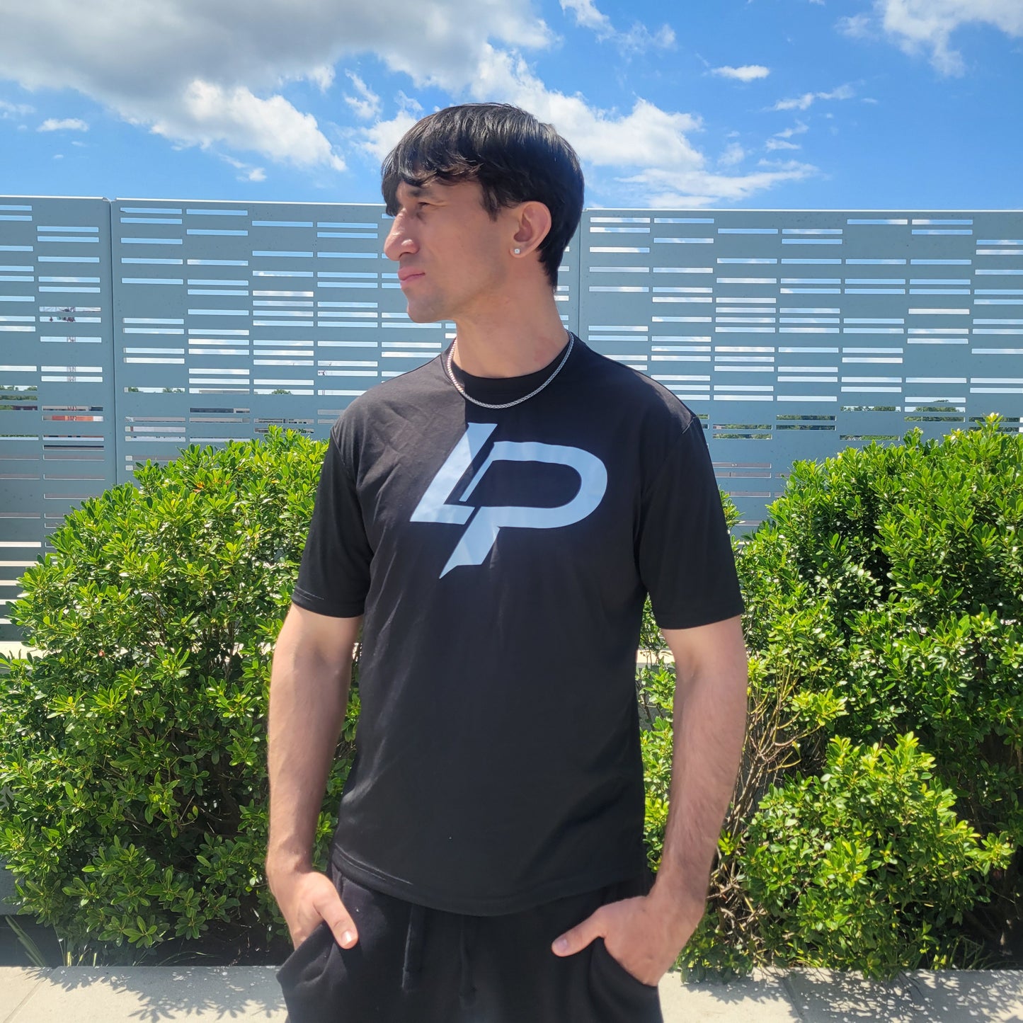 Man wearing a black unisex tee featuring a bold white "LP" monogram logo, photographed outdoors against green shrubs and a blue sky. Apparel lifestyle photo.