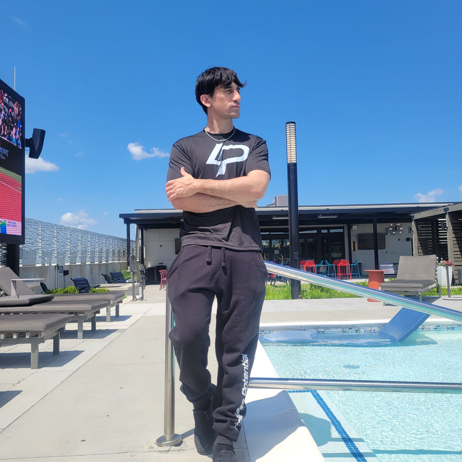 Man standing arms crossed by a rooftop pool wearing a black graphic tee and black joggers, sunny blue-sky backdrop, apparel lifestyle photo.