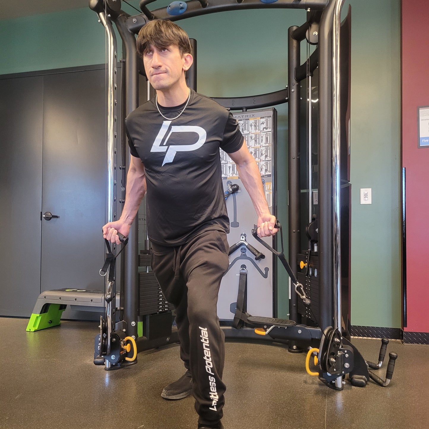 Man using a cable machine in a gym, wearing a black "LP" logo t-shirt and black "Limitless Potential" sweatpants, apparel lifestyle photo.