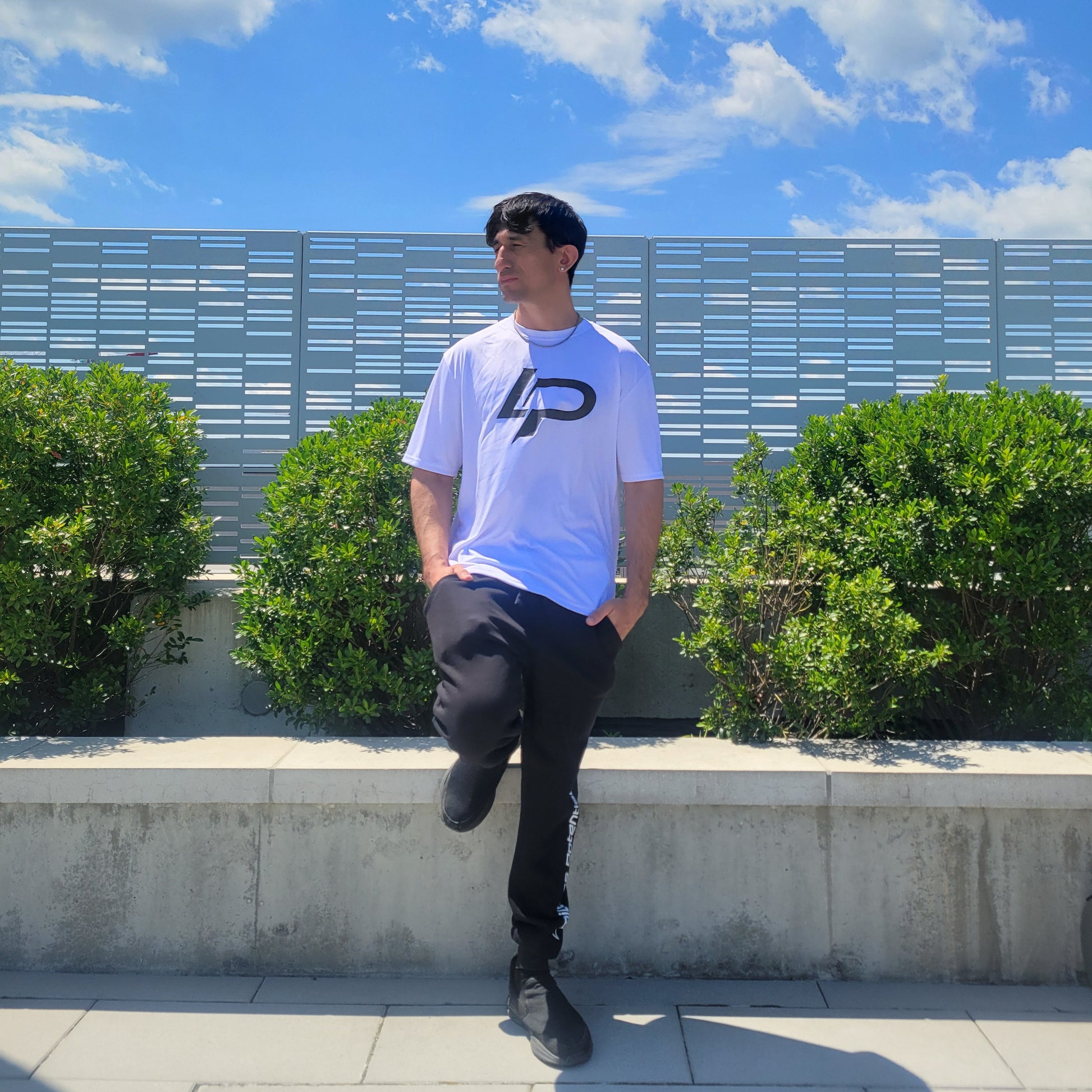 Male model wearing a white graphic tee with a bold black "LP" logo, paired with black joggers, posed outdoors against greenery and a blue sky; apparel lifestyle shot.
