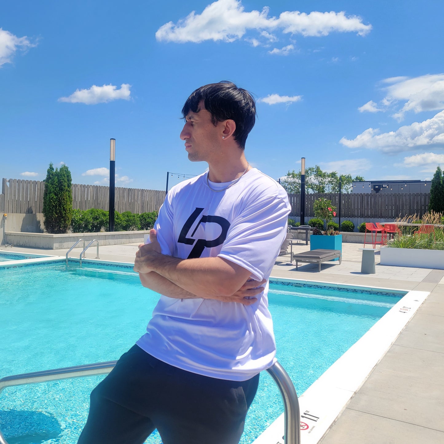 Man with arms crossed beside a rooftop pool wearing a white t-shirt with a bold black "LP" logo graphic, apparel lifestyle photo.