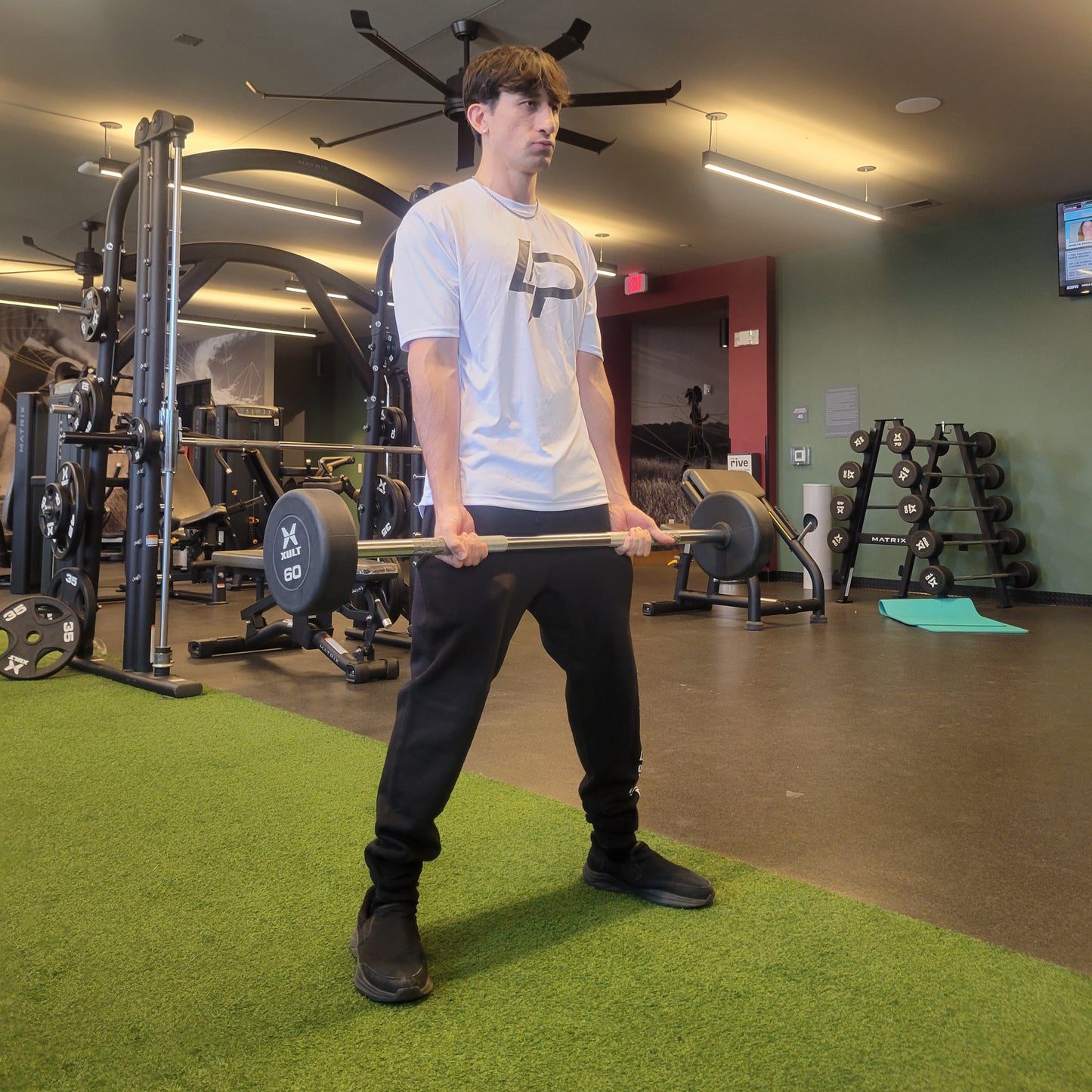 Man in a white graphic tee with bold black "LP" print on the chest holding a barbell in a modern gym, wearing black joggers; lifestyle apparel photo.