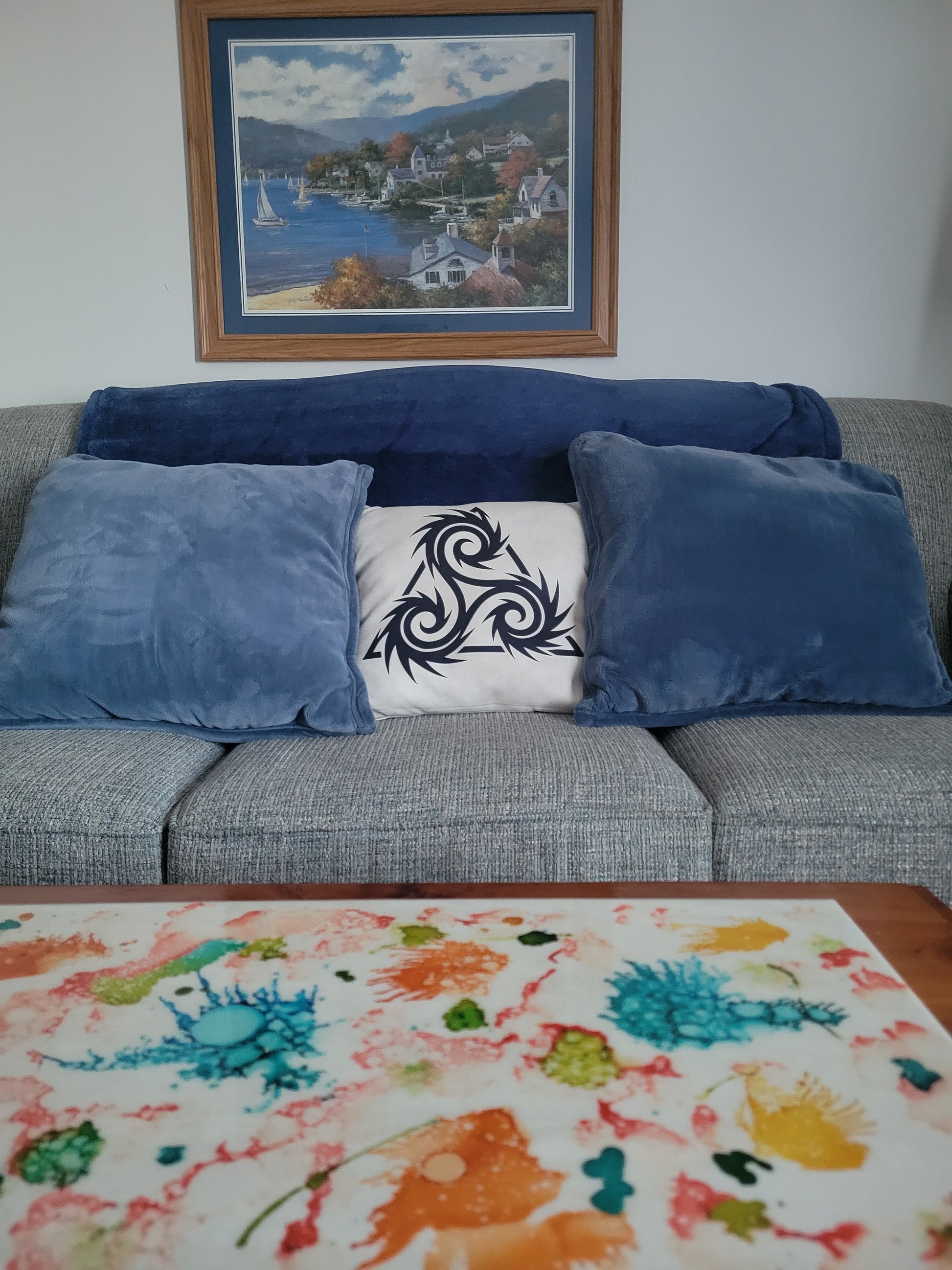 Beige throw pillow featuring a bold black Norse triskelion with sharp spiral arms, displayed on a gray sofa among blue velvet cushions — throw pillow.